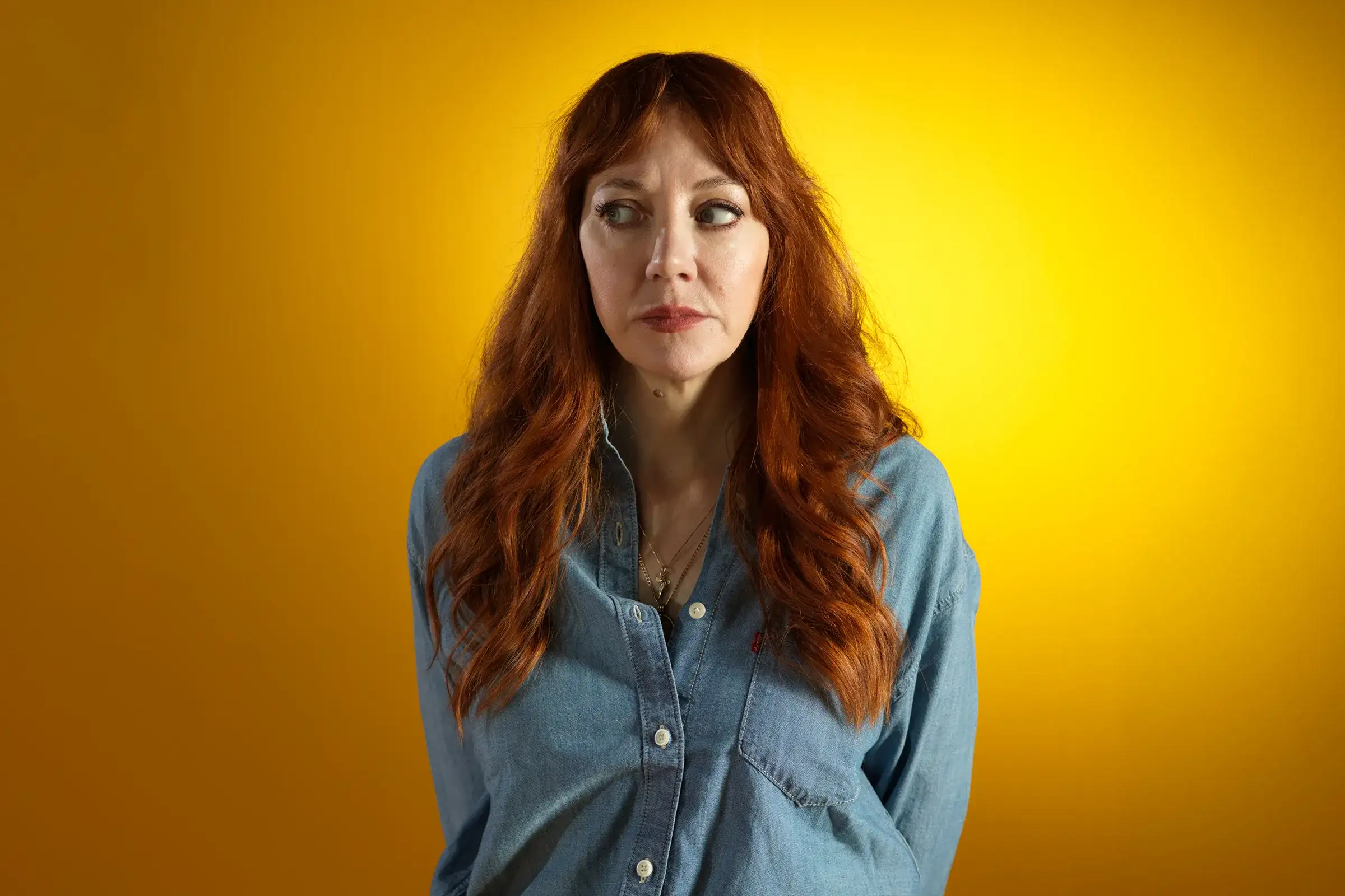 Diane Morgan stands against a yellow background, looking to one side with a serious expression.