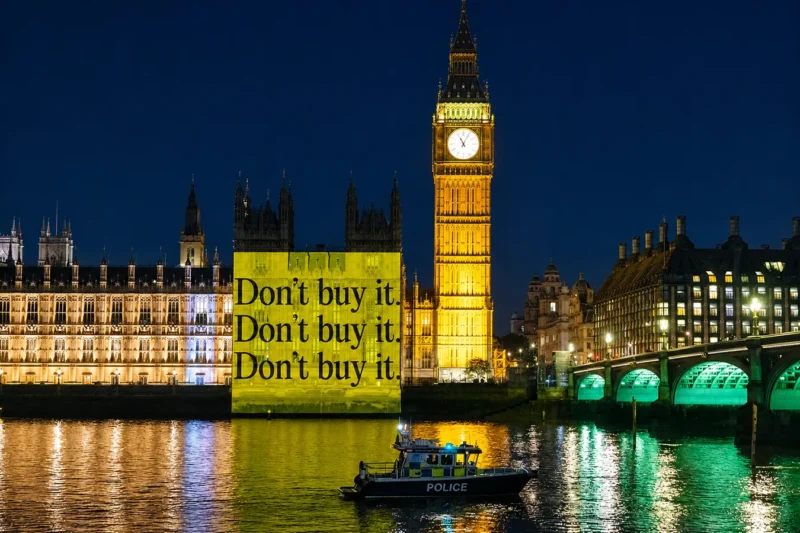 The Houses of Parliament lit up at night and against it is a projection of blaco text on a yellow background, which reads "Don't buy it." In the foreground is the Thames, and on it, in front of the projection, is a police boat.