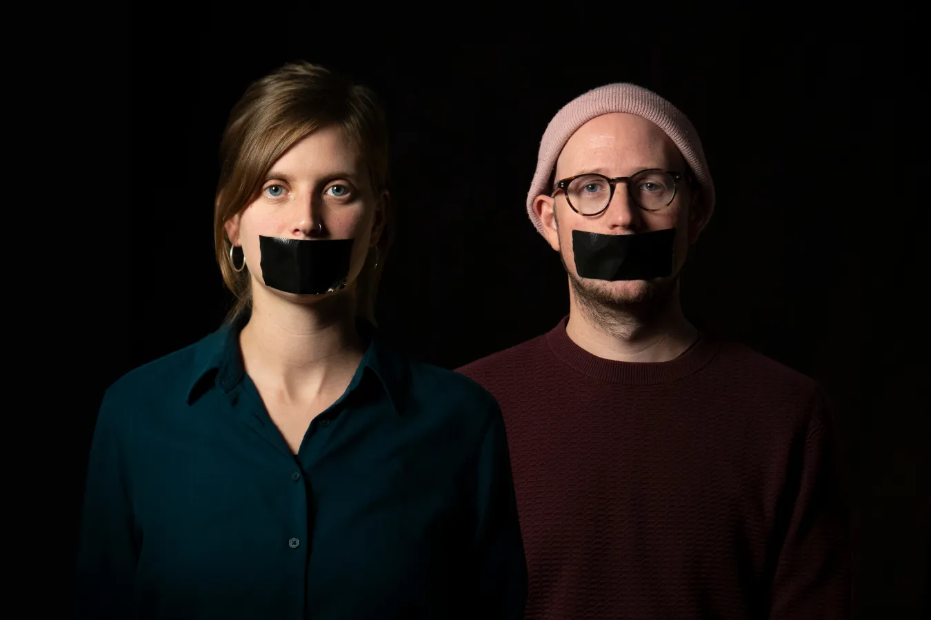 A woman and a man stand side by side against a black background, each with a strip of black tape over their mouth.