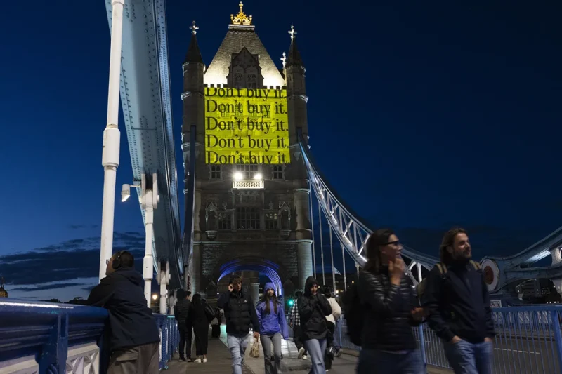 The iconic Tower Bridge, London, at night. People walking over the bridge and above them is a projection onto the tower, which reads “Don’t buy it”.