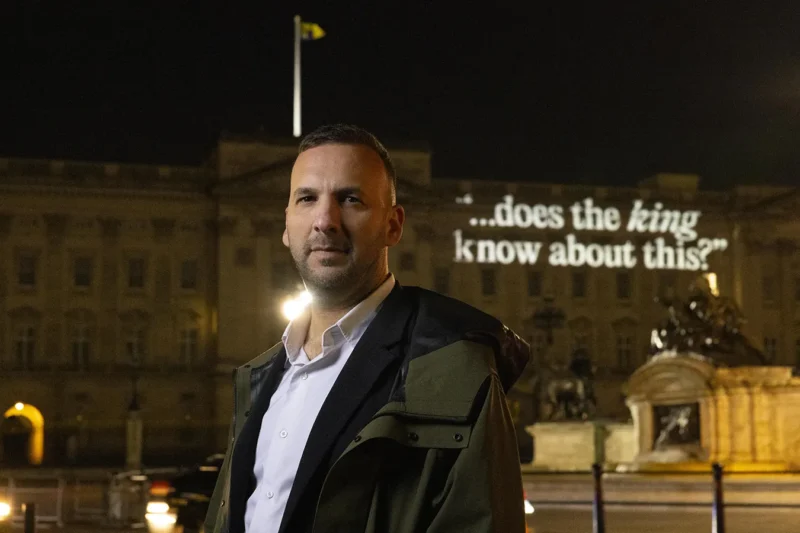 Leader of the Green Party stands in front of Buckingham Palace at night and onto the building, the words "Does the king know about this?" are projected.