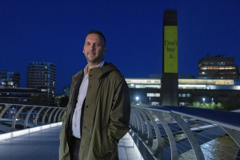 Leader of the Green Party Zack Polanski on Millennium Bridge, London at night, standing in front of a projection onto The Tate, which reads “Don’t buy it.”