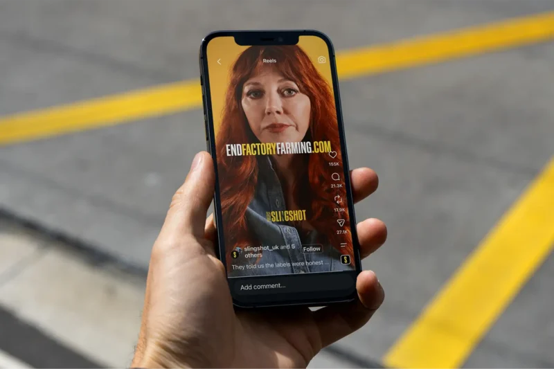 A hand holds a phone showing an End Factory Farming Instagram Reel featuring comedian Diane Morgan, with over 155,000 likes visible on screen.