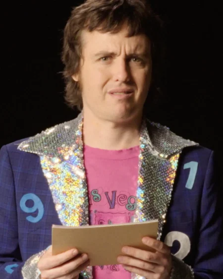 Comedian Sam Campbell holds a card while dressed as a quiz show host in a sequined jacket with numbers on it.