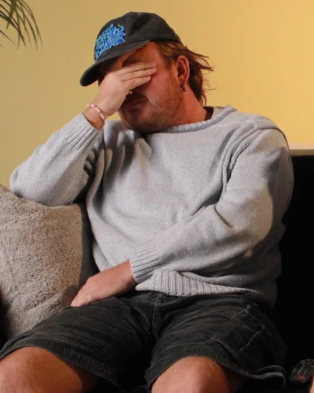 A man sitting on a sofa covers his face with his hand in a reaction of dismay.